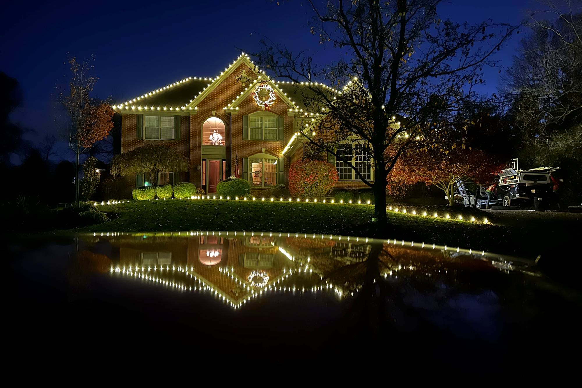 Brick two-story home with warm white Christmas lights on the roofline and a lit wreath, reflected in the wet driveway at dusk, installed by Liberty Christmas Lights