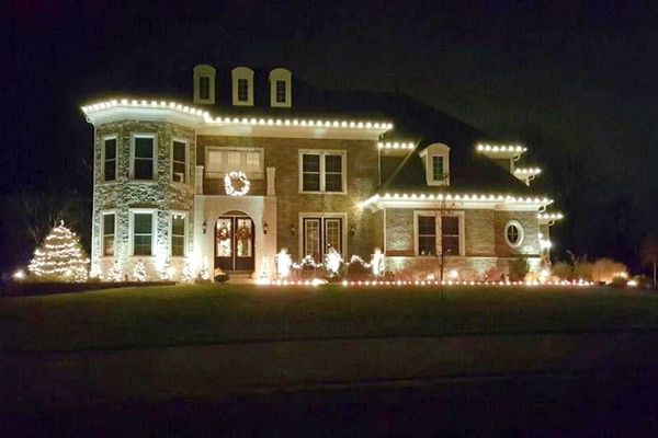 Brick and stone home with warm white roofline lights, a wreath, and a lit evergreen at the entry