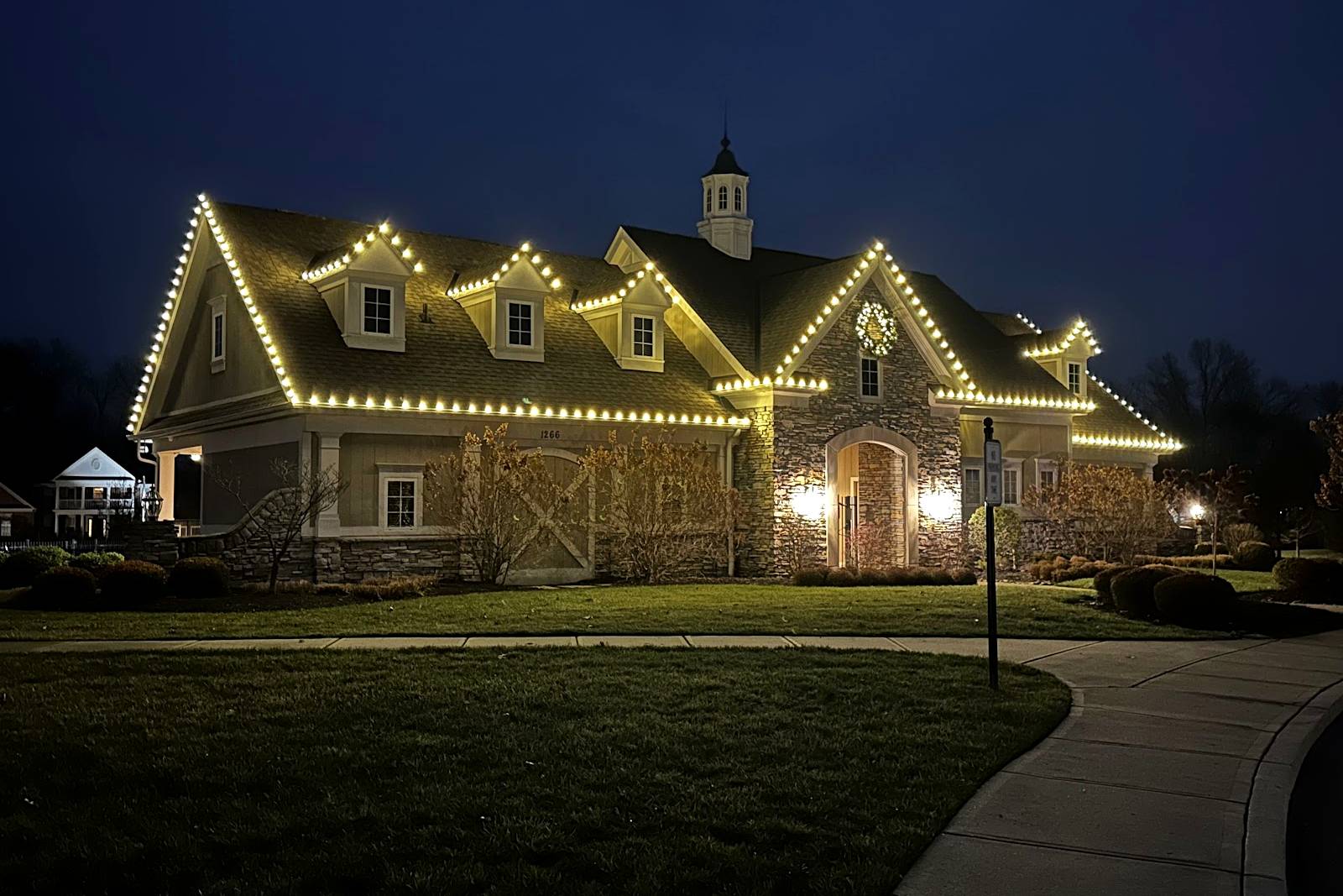 Stone and siding home with dormers and a cupola outlined in warm white Christmas lights at dusk, installed by Liberty Christmas Lights