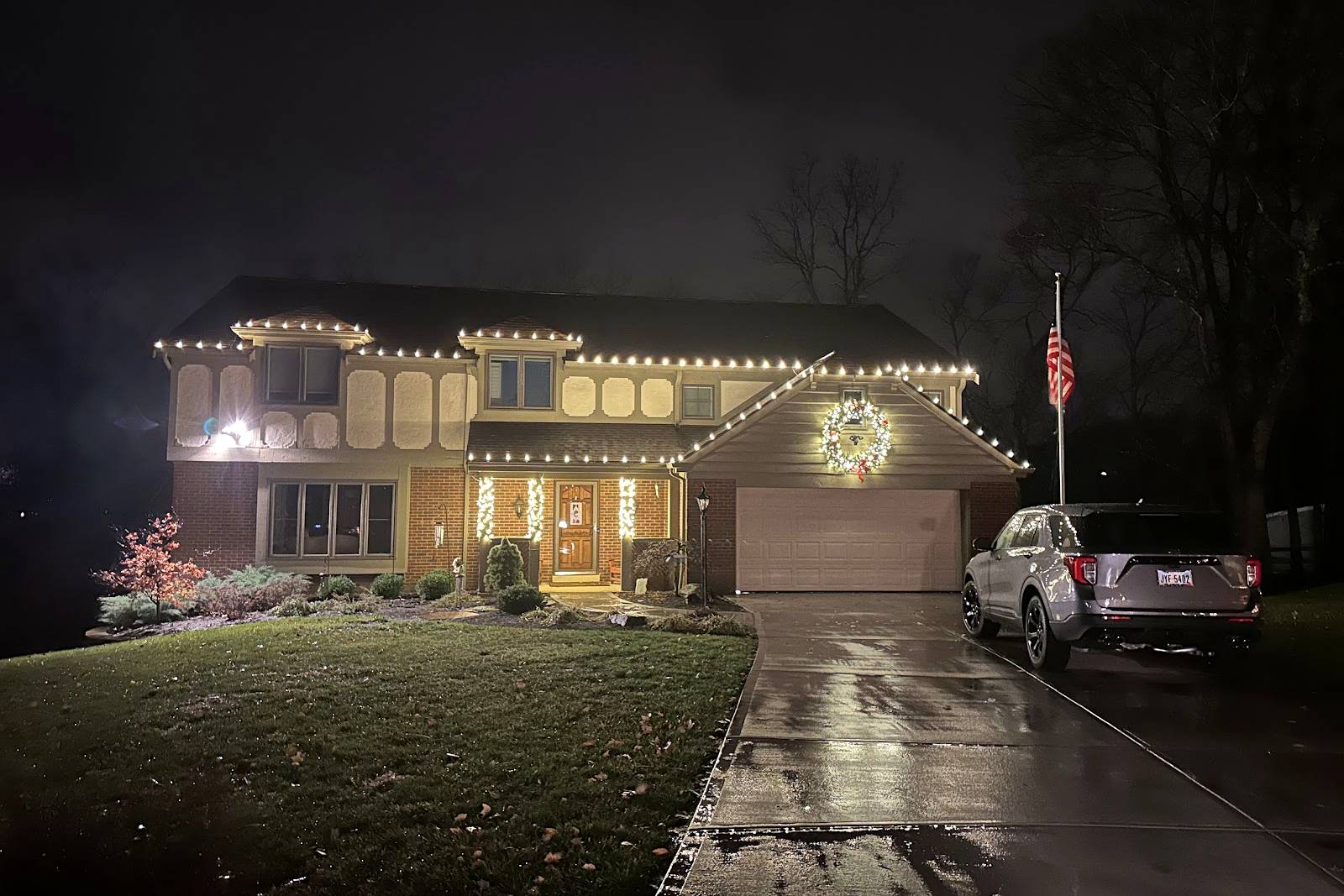 Two-story home with warm white roofline lights and a lit wreath over the garage, installed by Liberty Christmas Lights