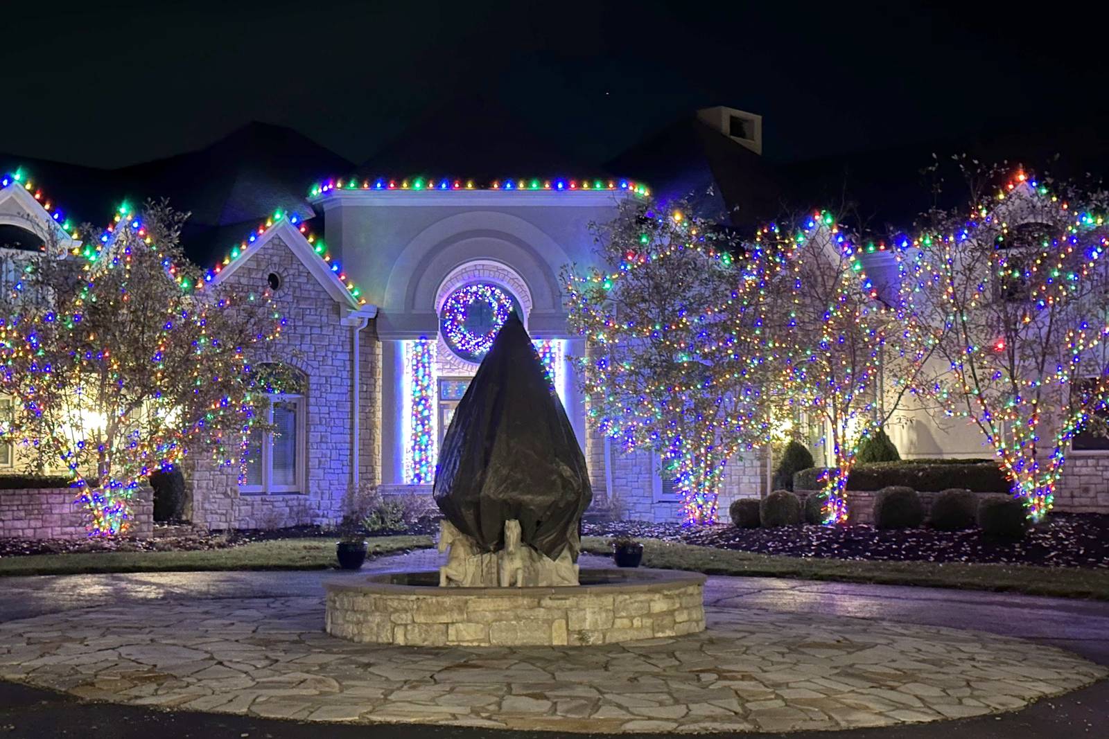 Large stone estate with multicolor roofline lights and lit trees around a circular drive, installed by Liberty Christmas Lights