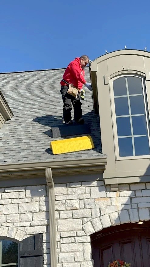 DJ working at the peak of a steep roof beside a stone dormer during a Christmas light install